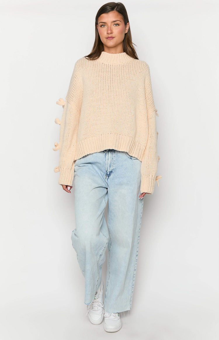 Front view of a cream chunky knit jumper with a mock neck and bow details along the long sleeves