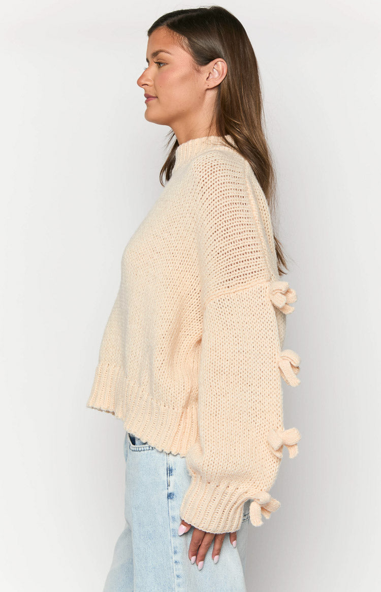 Side view of a cream knit jumper with mock neck, relaxed fit, and decorative bows down the long sleeves