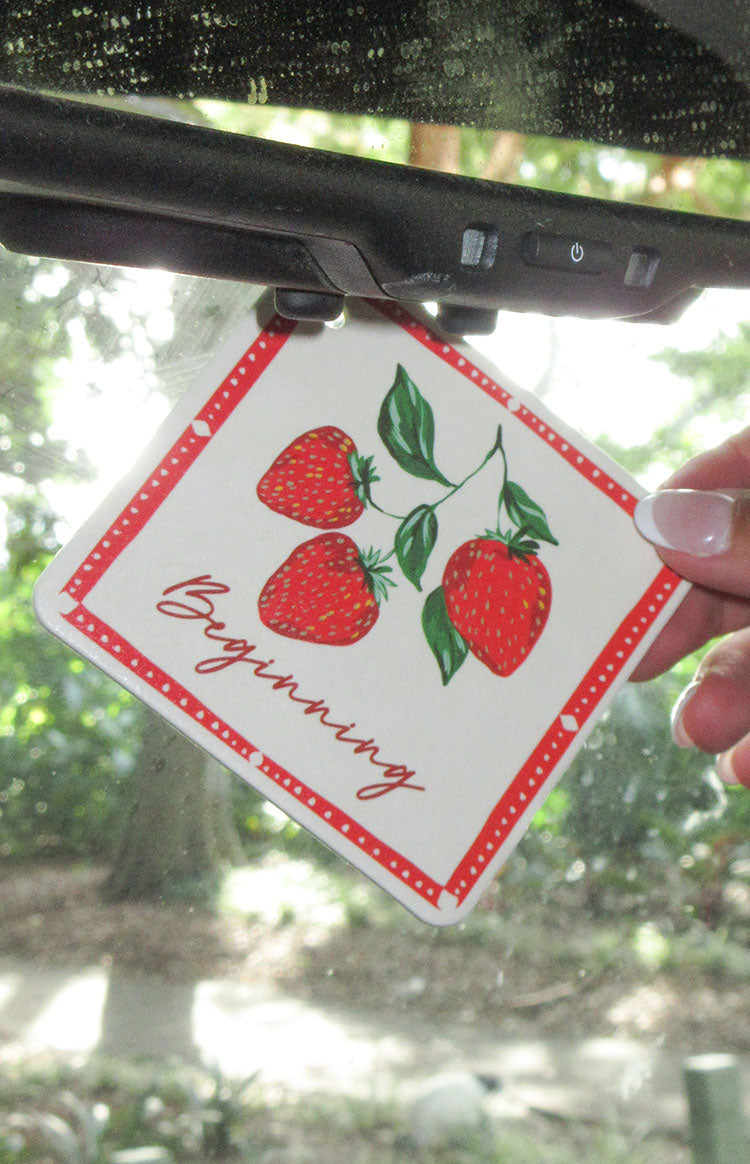 White square air freshener with a red dotted border and three red strawberries, hanging in a car