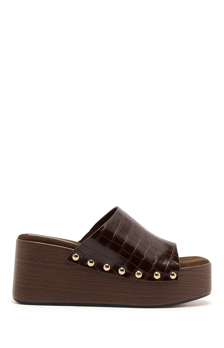 A single dark brown platform slide sandal features a wide strap with a glossy crocodile or alligator embossed faux leather finish. This strap is attached to the platform sole with a row of small brass studs. The sole is a chunky platform wedge with a dark wood-grain texture effect.