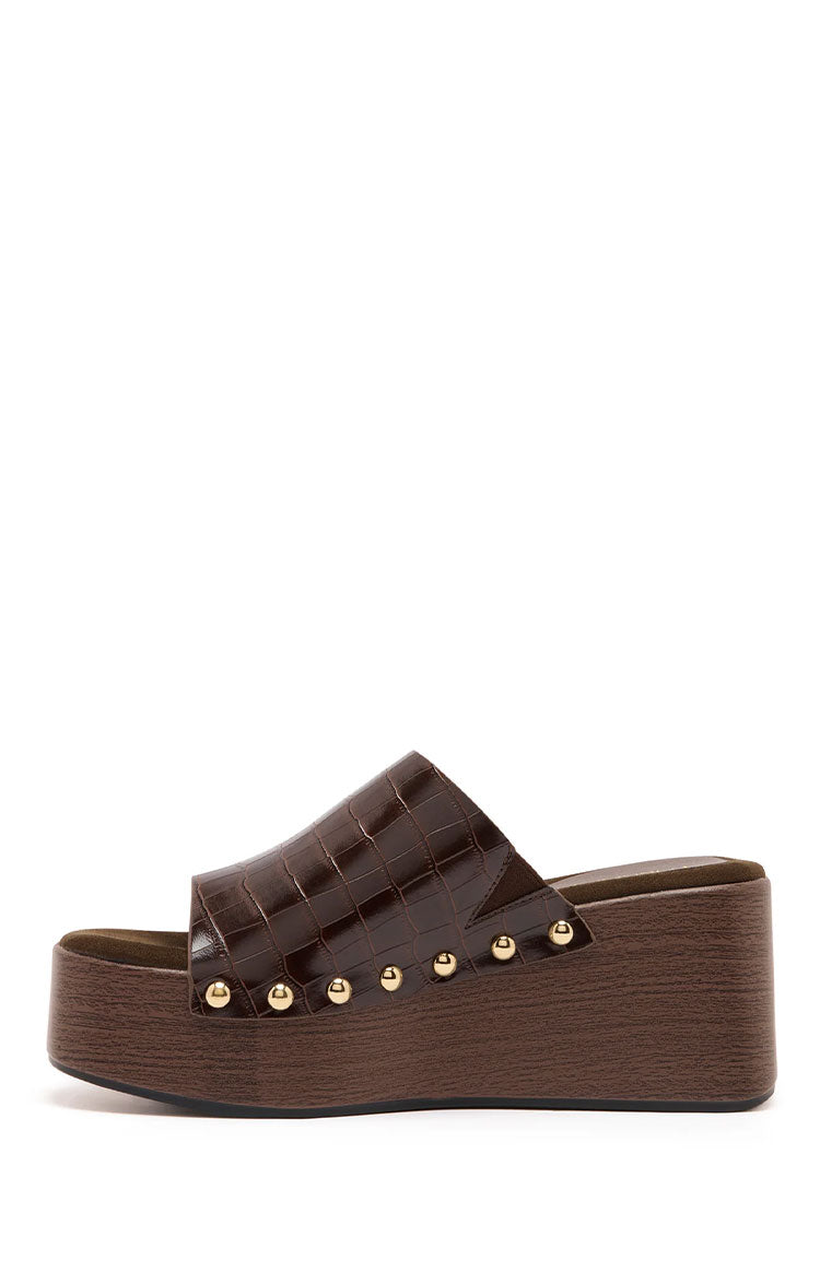 A side profile of a single dark brown platform wedge mule features a wide strap made of glossy crocodile-embossed faux leather. The strap is secured to the sole with a line of small gold-toned studs. The sole is a chunky platform wedge with a simulated dark wood-grain finish.
