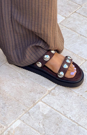 Close-up of a foot wearing a chocolate brown suede platform sandal with two straps adorned with large silver studs, paired with striped wide-leg pants.