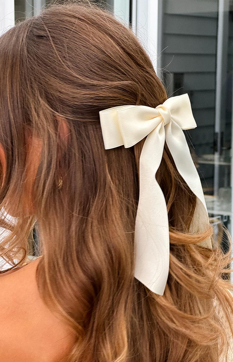 A cream satin bow hair clip with flowing tails, clipped into brown wavy hair.