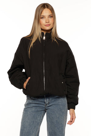 A close-up of our model wearing a black puffer bomber jacket with a relaxed slightly oversized fit. This jacket features a high stand collar and a prominent silver zipper down the front. It has an elasticized hem and cuffs with one hand tucked into a side pocket.