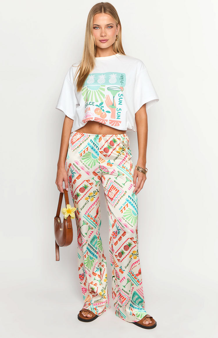 Full-length view of white wide-leg pants with a vibrant all-over print featuring pineapples, cocktails, and text