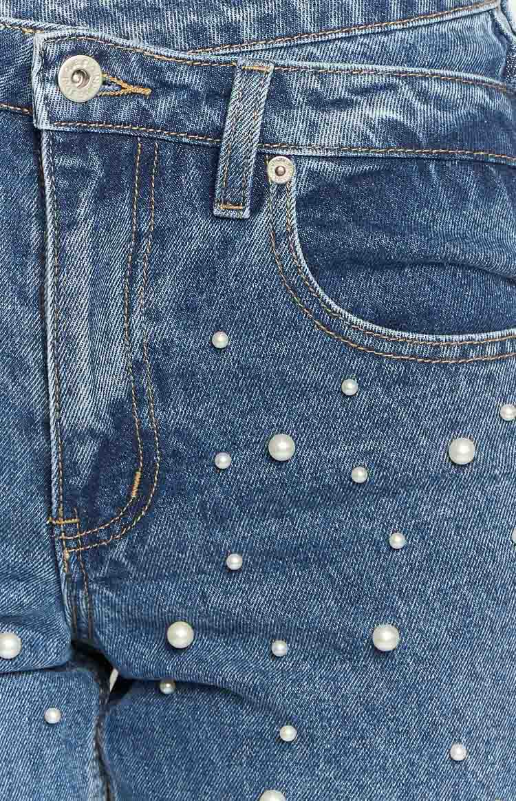 Close-up of the mid-wash denim jeans featuring a button closure, front pocket, and scattered pearl embellishments