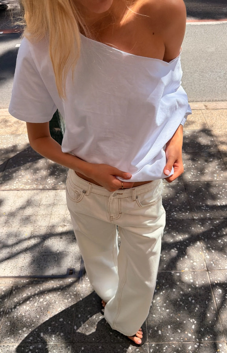 Model wearing a pair of white jeans featuring brown contrast stitching and a low rise fit. Styled with an off shoulder baggy tee, perfect for Fall!