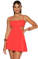 Our model wears a vibrant red mini-dress with a fit-and-flare silhouette. The dress features a sweetheart or gently curved neckline with delicate bobble trim and thin spaghetti straps. The most prominent design element is the geometric cutouts at the waist and side trimmed with a matching bobble detail. The skirt is plain and flares out.