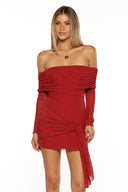 A half body shot of a woman wearing a tight short red dress made of ruched mesh fabric. The dress features an off the shoulder neckline long sheer sleeves and a draped panel of fabric tied in a knot at the hip creating an asymmetric hemline and detail.