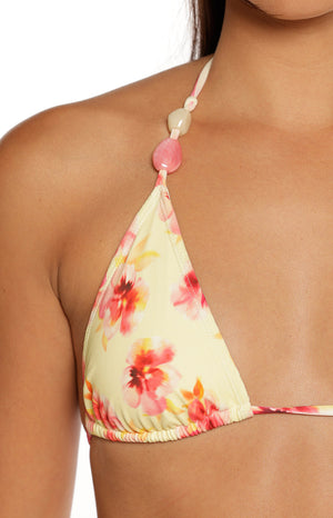 A very tight close-up focuses on the bikini top of a triangle bikini cup. The swimwear fabric is creamy yellow with a vibrant pink and red floral print. The thin halter strap is decorated with two large beads: a round bright pink stone and an elongated pale translucent bead positioned just above the pink one.