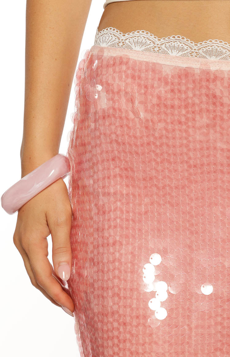 An extreme close up detail shot of the waistline of a pink sequin skirt. The image focuses on the texture of the pink sequins and the delicate scalloped white lace trim bordering the top edge of the skirt.
