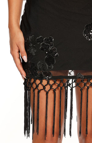 A close-up focusing on the hemline of a black mini-dress. The sheer fabric of the skirt is decorated with a sequined black floral appliqué. The bottom edge of the dress is finished with a band of macrame-style knotting from which long vertical black fringe tassels hang down over the thigh.