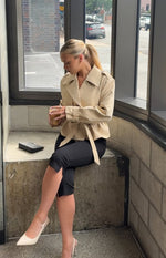 Side-front view of a cream cropped trench coat with a wide collar, shoulder epaulets, and a tied waist belt